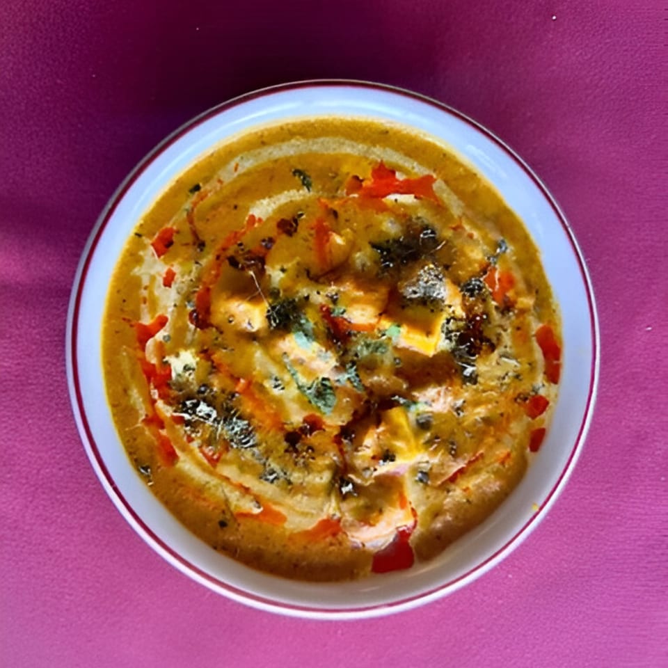 A bowl of creamy Indian paneer butter masala, garnished with herbs and red oil swirls.