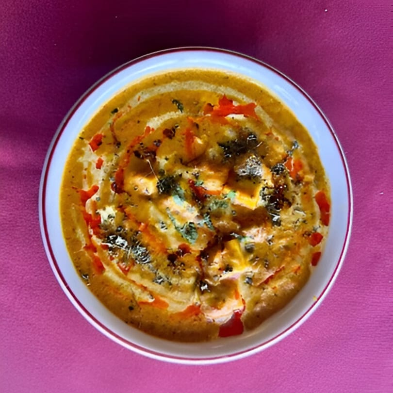 A bowl of creamy Indian paneer butter masala, garnished with herbs and red oil swirls.