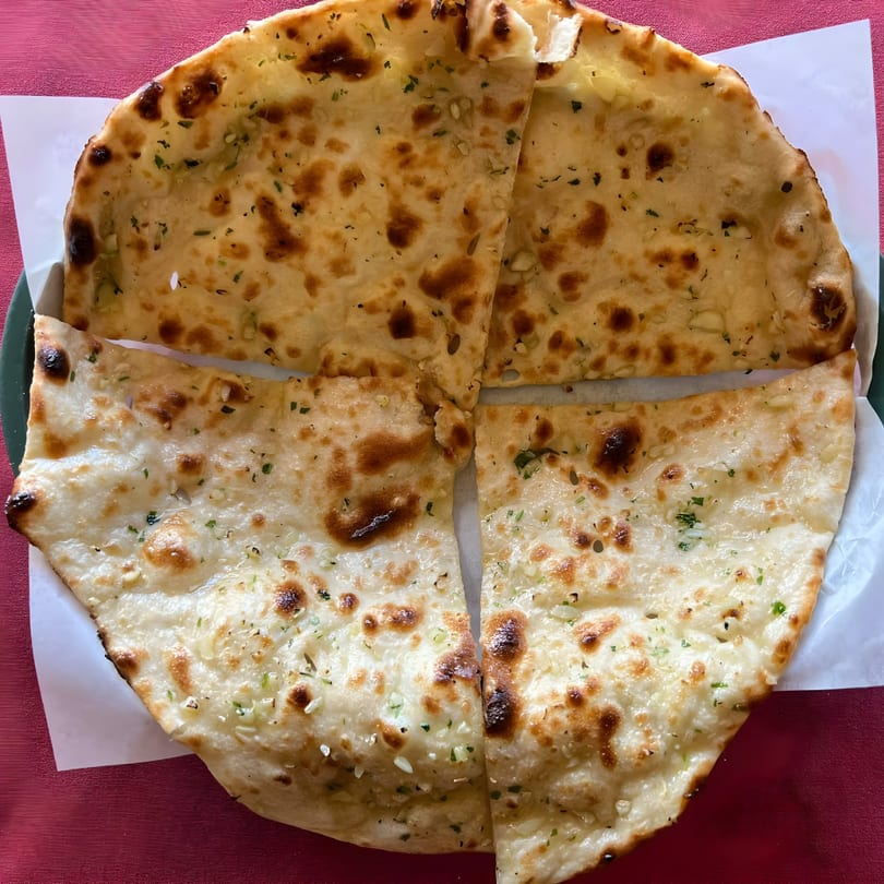 Four pieces of garlic naan bread, golden brown and garnished with herbs, on a white paper liner.